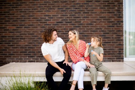 Téléchargez les photos : Famille avec une mère, un père et une fille assis dehors sur les marches d'un porche d'une maison en brique - en image libre de droit