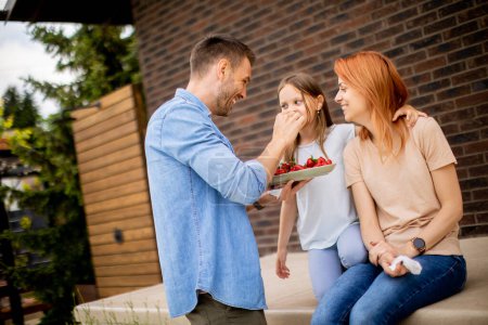 Téléchargez les photos : Family with a mother, father and daughter sitting outside on a steps of a front porch of a brick house and eating strawberries - en image libre de droit