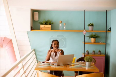Foto de Mujer negra bastante joven bebiendo café mientras trabaja en el portátil en el café - Imagen libre de derechos