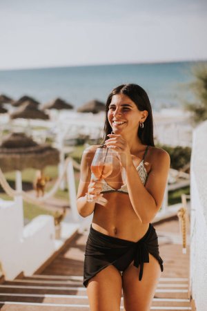 Téléchargez les photos : Souriant jeune femme en bikini Profiter de vacances sur la plage tout en buvant un cocktail - en image libre de droit