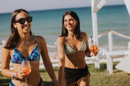 Téléchargez les photos : Deux jeunes femmes souriantes en bikini profitant de vacances sur la plage tout en buvant un cocktail - en image libre de droit