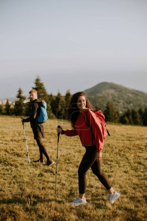 Photo for Smiling young couple walking with backpacks over green hills - Royalty Free Image