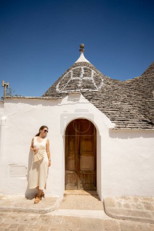 Téléchargez les photos : Une jeune femme vêtue d'une robe blanche et d'un chapeau se relaxant sur des étoiles par une journée ensoleillée lors d'une visite touristique à Alberobello, en Italie - en image libre de droit