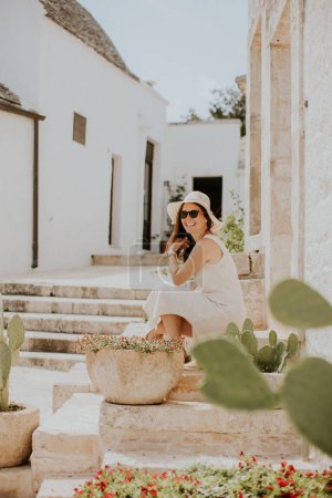 Téléchargez les photos : Une jeune femme en robe blanche et chapeau relaxant sur les escaliers par une journée ensoleillée lors d'une visite touristique à Alberobello, Italie - en image libre de droit