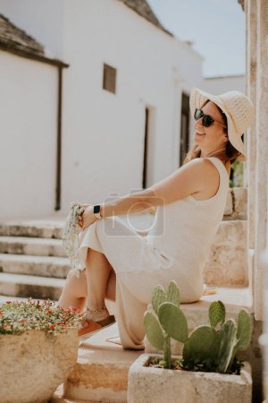Téléchargez les photos : Une jeune femme en robe blanche et chapeau relaxant sur les escaliers par une journée ensoleillée lors d'une visite touristique à Alberobello, Italie - en image libre de droit