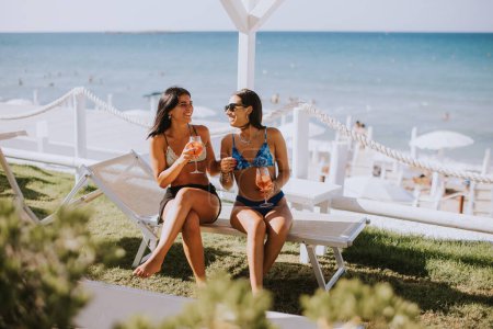 Foto de Dos jóvenes sonrientes en bikini disfrutando de vacaciones en la playa mientras beben cóctel - Imagen libre de derechos