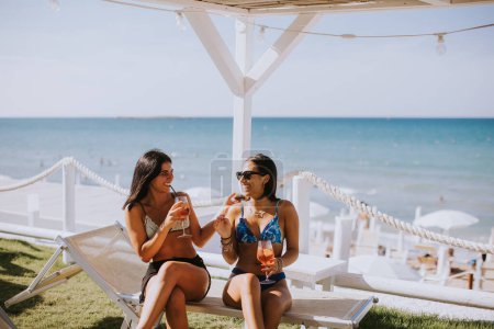 Foto de Dos jóvenes sonrientes en bikini disfrutando de vacaciones en la playa mientras beben cóctel - Imagen libre de derechos