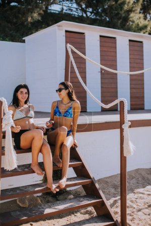 Téléchargez les photos : Deux jeunes femmes souriantes en bikini assis et profitant de vacances sur la plage - en image libre de droit
