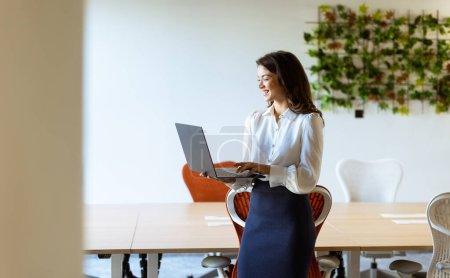 Téléchargez les photos : Jeune femme d'affaires avec ordinateur portable debout dans le bureau moderne - en image libre de droit