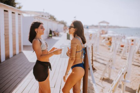 Téléchargez les photos : Deux jeunes femmes souriantes en bikini debout près des cabines et profitant de vacances sur la plage - en image libre de droit