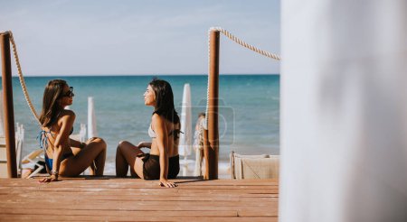 Foto de Dos jóvenes sonrientes en bikini sentadas y disfrutando de unas vacaciones en la playa - Imagen libre de derechos