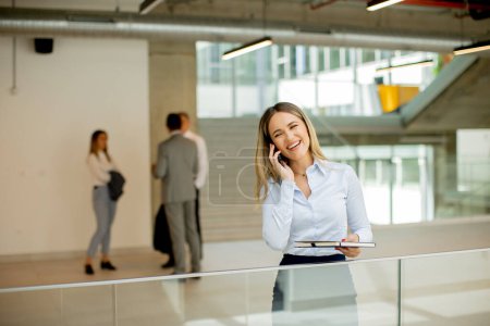 Téléchargez les photos : Jeune femme d'affaires avec ordinateur portable en utilisant le téléphone mobile et debout dans le mur de bureau moderne - en image libre de droit