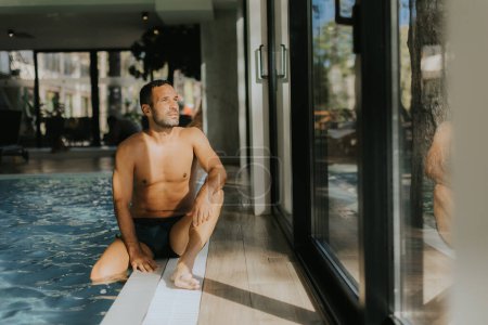Téléchargez les photos : Beau jeune homme relaxant au bord d'une piscine intérieure - en image libre de droit