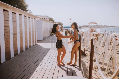 Téléchargez les photos : Deux jeunes femmes souriantes en bikini debout près des cabines et profitant de vacances sur la plage - en image libre de droit