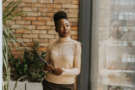 Téléchargez les photos : Une jolie jeune femme d'affaires afro-américaine avec tablette numérique debout près du mur de briques dans le bureau de style industriel - en image libre de droit