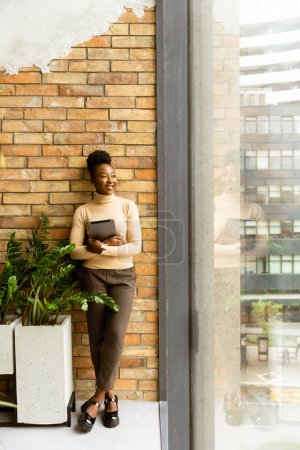 Photo for One pretty young African American business woman with digital tablet standing by the brick wall in the industrial style office - Royalty Free Image