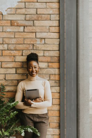 Photo for One pretty young African American business woman with digital tablet standing by the brick wall in the industrial style office - Royalty Free Image