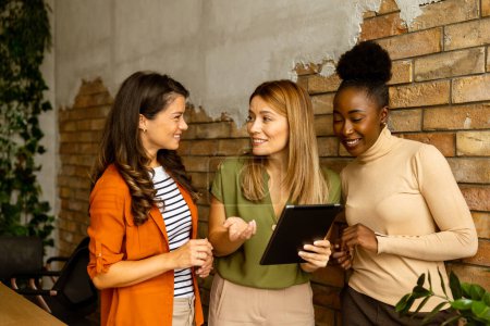 Téléchargez les photos : Trois jolies jeunes femmes d'affaires avec tablette numérique debout près du mur de briques dans le bureau de style industriel - en image libre de droit