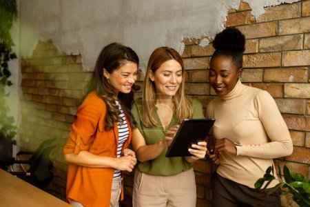 Foto de Tres mujeres de negocios bastante jóvenes con tableta digital de pie junto a la pared de ladrillo en la oficina de estilo industrial - Imagen libre de derechos