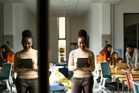 Foto de Joven mujer afroamericana de pie con tableta digital frente a su equipo en la oficina moderna - Imagen libre de derechos