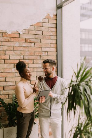 Téléchargez les photos : Beau jeune couple d'affaires multiethnique avec tablette numérique debout et discuter dans le bureau de style industriel - en image libre de droit