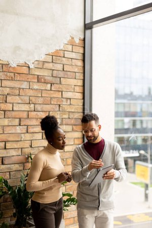 Téléchargez les photos : Beau jeune couple d'affaires multiethnique avec tablette numérique debout et discuter dans le bureau moderne - en image libre de droit