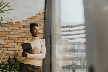 Foto de Una mujer de negocios afroamericana bastante joven con tableta digital de pie junto a la pared de ladrillo en la oficina de estilo industrial - Imagen libre de derechos