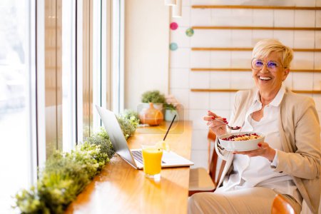 Photo for Good looking senior woman working on a laptop, drinking orange juice and having a healthy breakfast in cafeteria - Royalty Free Image