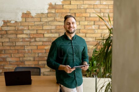 Cheerful man stands confidently holding a digital tablet in a contemporary office space with an exposed brick wall, symbolizing a blend of modern tech and rustic charm