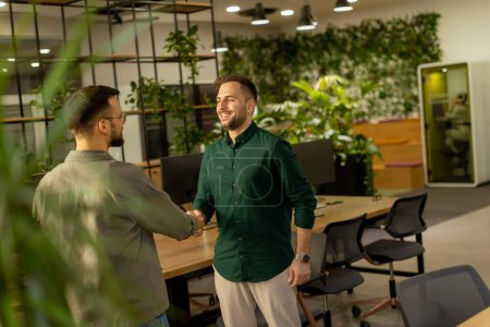 Dos profesionales intercambian un cálido apretón de manos en el acogedor ambiente de un espacio de oficina adornado con zonas verdes, lo que indica una colaboración exitosa a medida que el día termina.