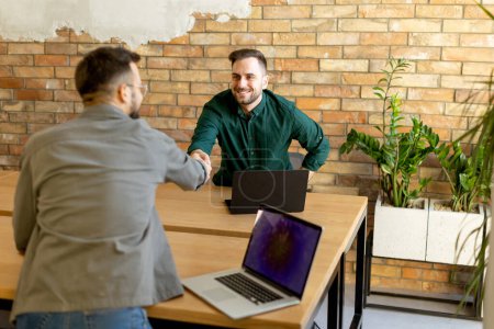 Photo for Two professionals engage in a welcoming handshake across a wooden table adorned with laptops, signaling a successful meeting or partnership in a contemporary brick-walled office - Royalty Free Image