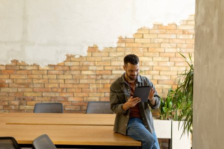 Cheerful man stands confidently holding a digital tablet in a contemporary office space with an exposed brick wall, symbolizing a blend of modern tech and rustic charm