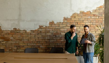 Foto de Dos profesionales se dedican a una conversación enfocada mientras sostienen la tableta digital en un espacio de oficina contemporáneo, con ladrillos expuestos rústicos y luz natural cálida - Imagen libre de derechos
