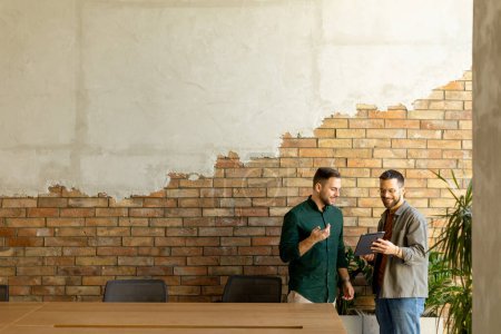 Photo for Two professionals are engaged in a focused conversation while holding digital tablet in a contemporary office space, featuring rustic exposed brickwork and warm natural light - Royalty Free Image