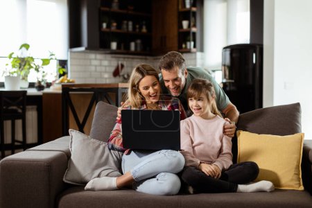 Foto de Familia alegre de tres pasa tiempo de calidad juntos en el sofá de la sala de estar, compartiendo un momento alrededor de un ordenador portátil en su hogar cómodo - Imagen libre de derechos