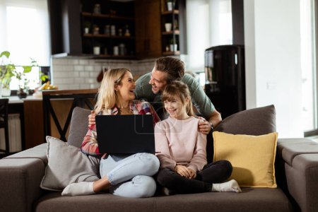 Foto de Familia alegre de tres pasa tiempo de calidad juntos en el sofá de la sala de estar, compartiendo un momento alrededor de un ordenador portátil en su hogar cómodo - Imagen libre de derechos