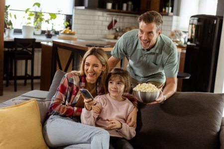 Una familia de tres miembros está cómodamente enclavada en un sofá, sus rostros reflejan emoción y atención mientras comparten un tazón de palomitas de maíz durante una noche de cine suspendida.