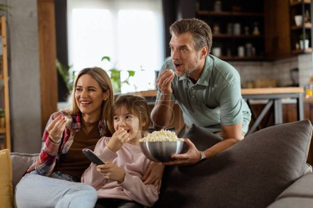 Eine dreiköpfige Familie liegt gemütlich auf einer Couch, ihre Gesichter spiegeln Aufregung und Aufmerksamkeit wider, während sie sich während eines spannenden Filmabends eine Schüssel Popcorn teilen.