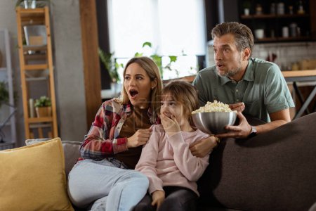 A family of three is comfortably nestled on a couch, their faces reflecting excitement and attentiveness as they share a bowl of popcorn during a suspenseful movie night