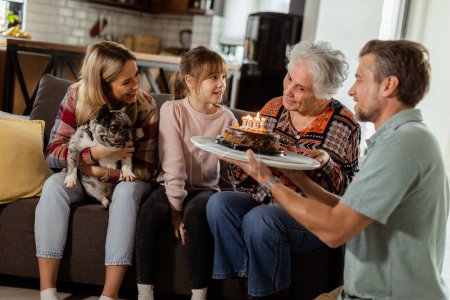 Photo for Heartwarming scene unfolds as a multi-generational family gathers on a couch to present a birthday cake to a delighted grandmother, creating memories to cherish - Royalty Free Image