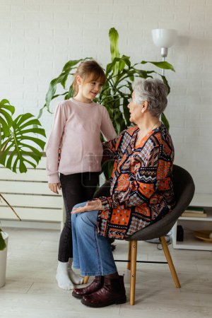 Foto de En una habitación suavemente iluminada, una joven se encuentra junto a una planta en maceta, compartiendo un momento de conexión y conversación con su abuela anciana, la alegría y el calor entre ellos palpable - Imagen libre de derechos