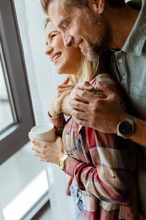 Photo for In a softly lit room, a man lovingly embraces a woman from behind as they both gaze out the window, sharing a quiet, intimate moment together - Royalty Free Image
