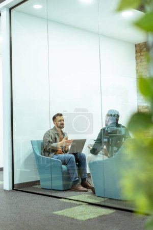 Two professionals engage in a conversation while seated comfortably in an office lounge, surrounded by greenery and contemporary design elements