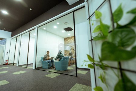 Foto de Dos profesionales conversan sentados cómodamente en un salón de oficina, rodeado de vegetación y elementos de diseño contemporáneo - Imagen libre de derechos