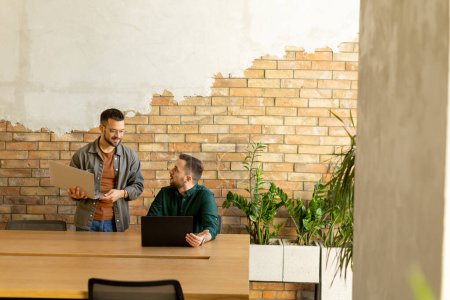 Photo for Two smiling professionals engage in a collaborative work session at a wooden table, their camaraderie evident in a contemporary office setting with an exposed brick wall backdrop - Royalty Free Image