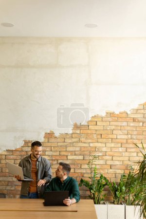 Foto de Dos profesionales sonrientes participan en una sesión de trabajo colaborativo en una mesa de madera, su camaradería evidente en un entorno de oficina contemporáneo con un fondo de pared de ladrillo visto - Imagen libre de derechos
