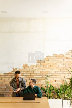 Zwei lächelnde Profis nehmen an einem Holztisch an einer gemeinsamen Arbeitssitzung teil, deren Kameradschaft in einem modernen Büroambiente vor einer exponierten Backsteinwand deutlich wird.