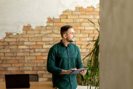 Photo for Cheerful man stands confidently holding a digital tablet in a contemporary office space with an exposed brick wall, symbolizing a blend of modern tech and rustic charm - Royalty Free Image