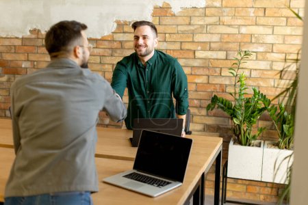 Photo for Two professionals engage in a welcoming handshake across a wooden table adorned with laptops, signaling a successful meeting or partnership in a contemporary brick-walled office - Royalty Free Image