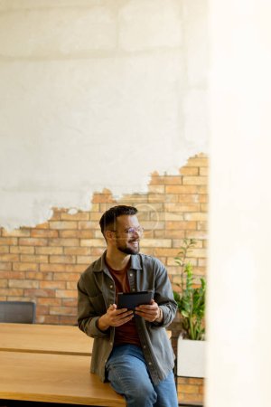 Hombre alegre se para con confianza sosteniendo una tableta digital en un espacio de oficina contemporáneo con una pared de ladrillo visto, que simboliza una mezcla de tecnología moderna y encanto rústico